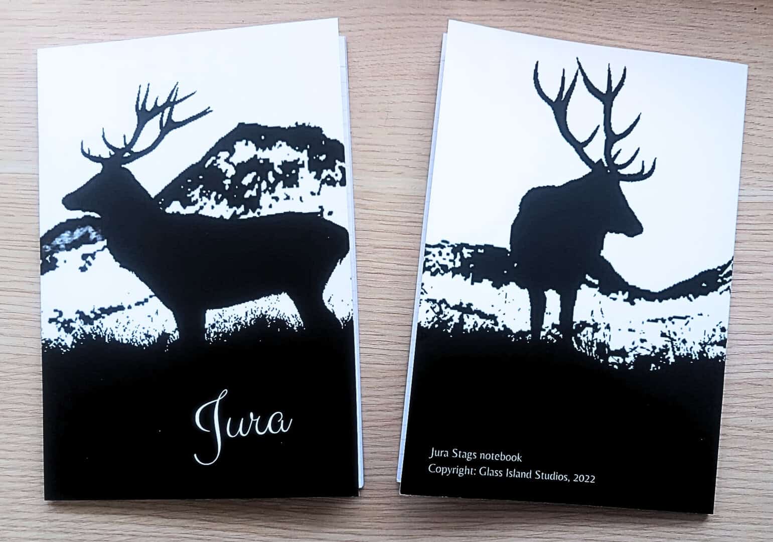 Photo of two notebooks of the same design featuring a black and white artist impression of two stags silhouetted against mountains. One notebook is face up and the other is face down.